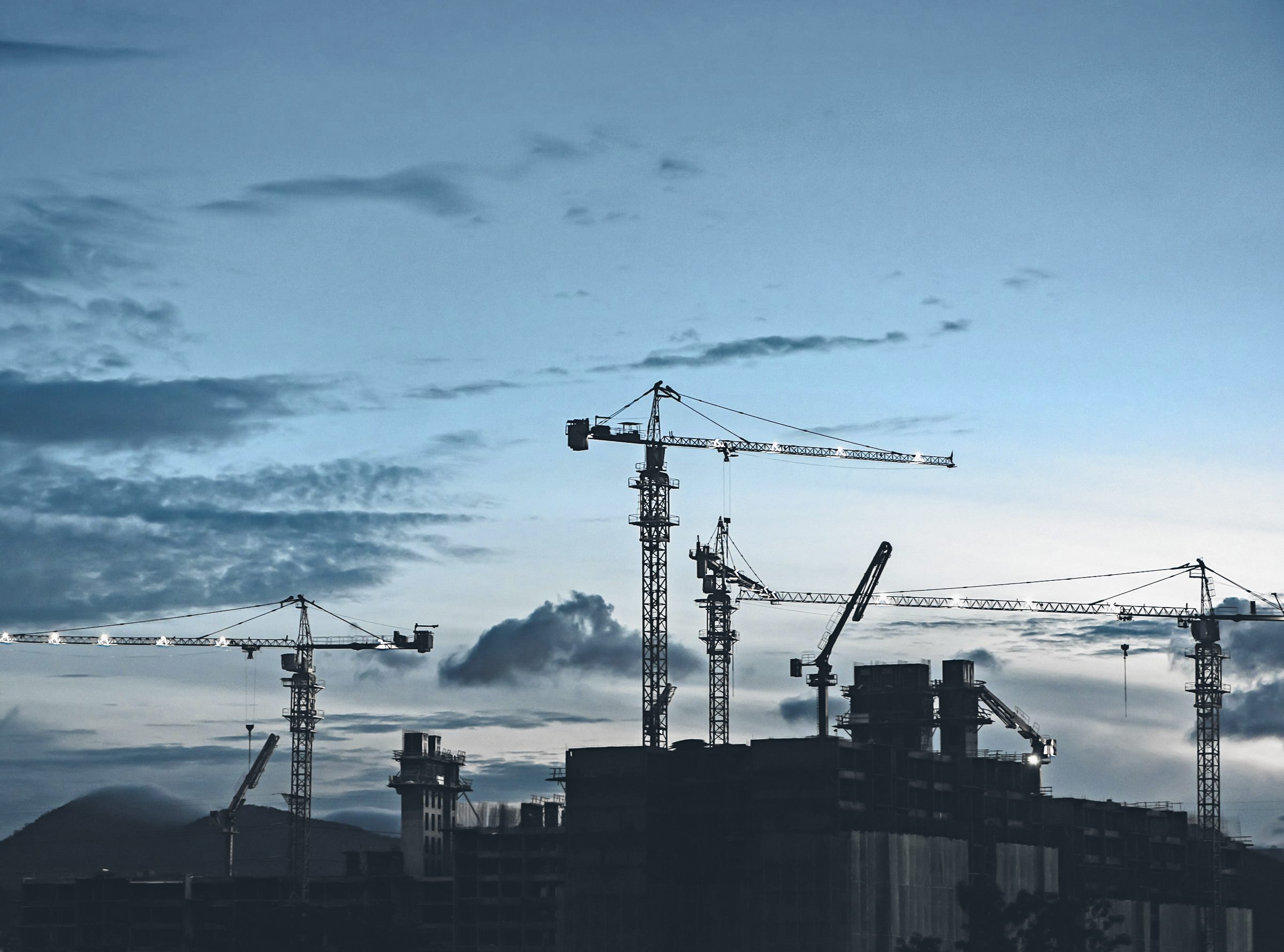Tower cranes standing over the frames of high-rise buildings at dusk, under a cloud-streaked blue sky.