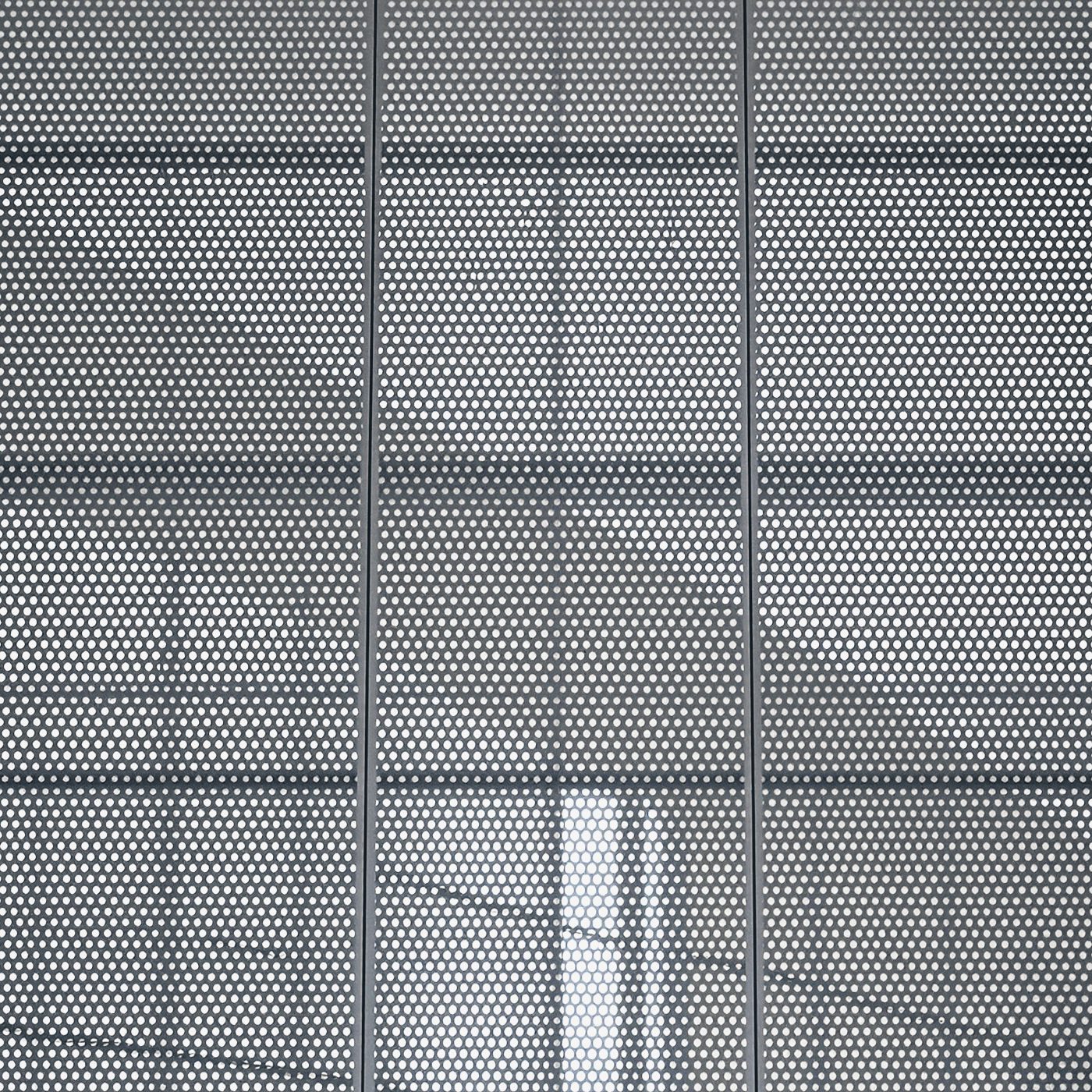 Close-up of perforated metal facade panels with folded edges in front of glazing.
