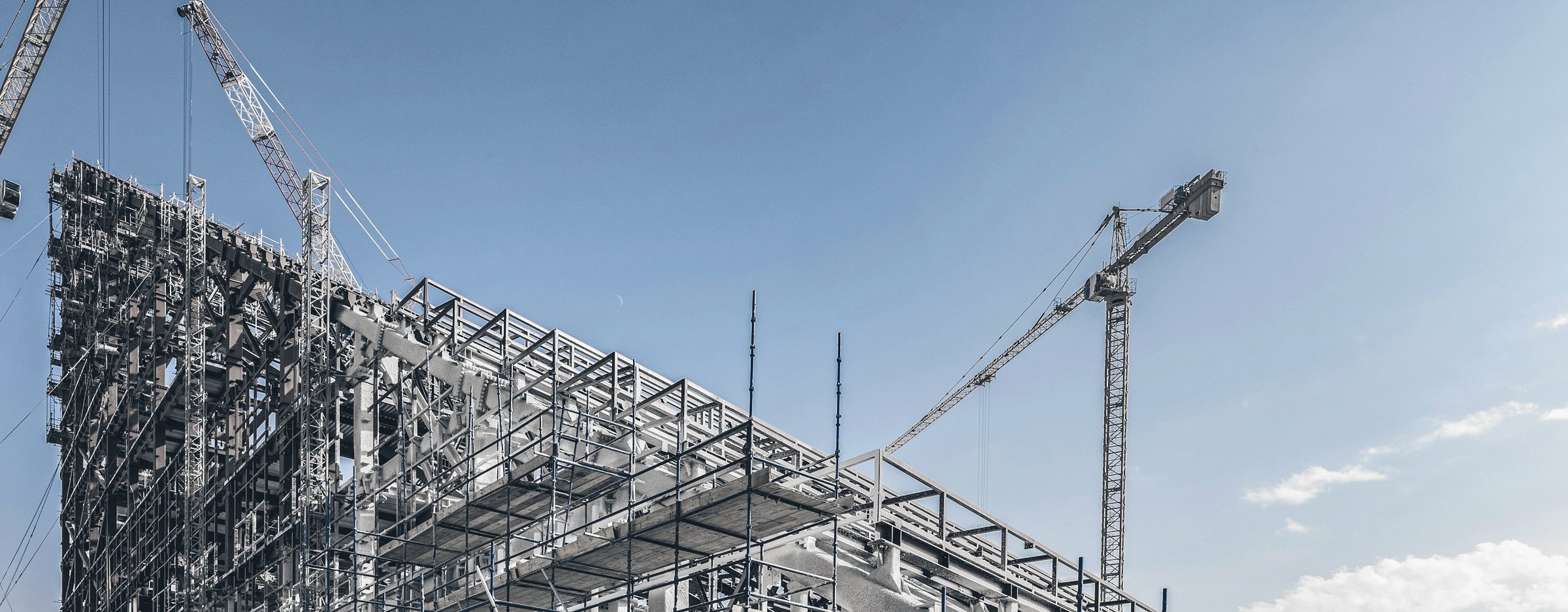 A large steel frame under erection, wrapped in scaffolding, with tower cranes against a blue sky.