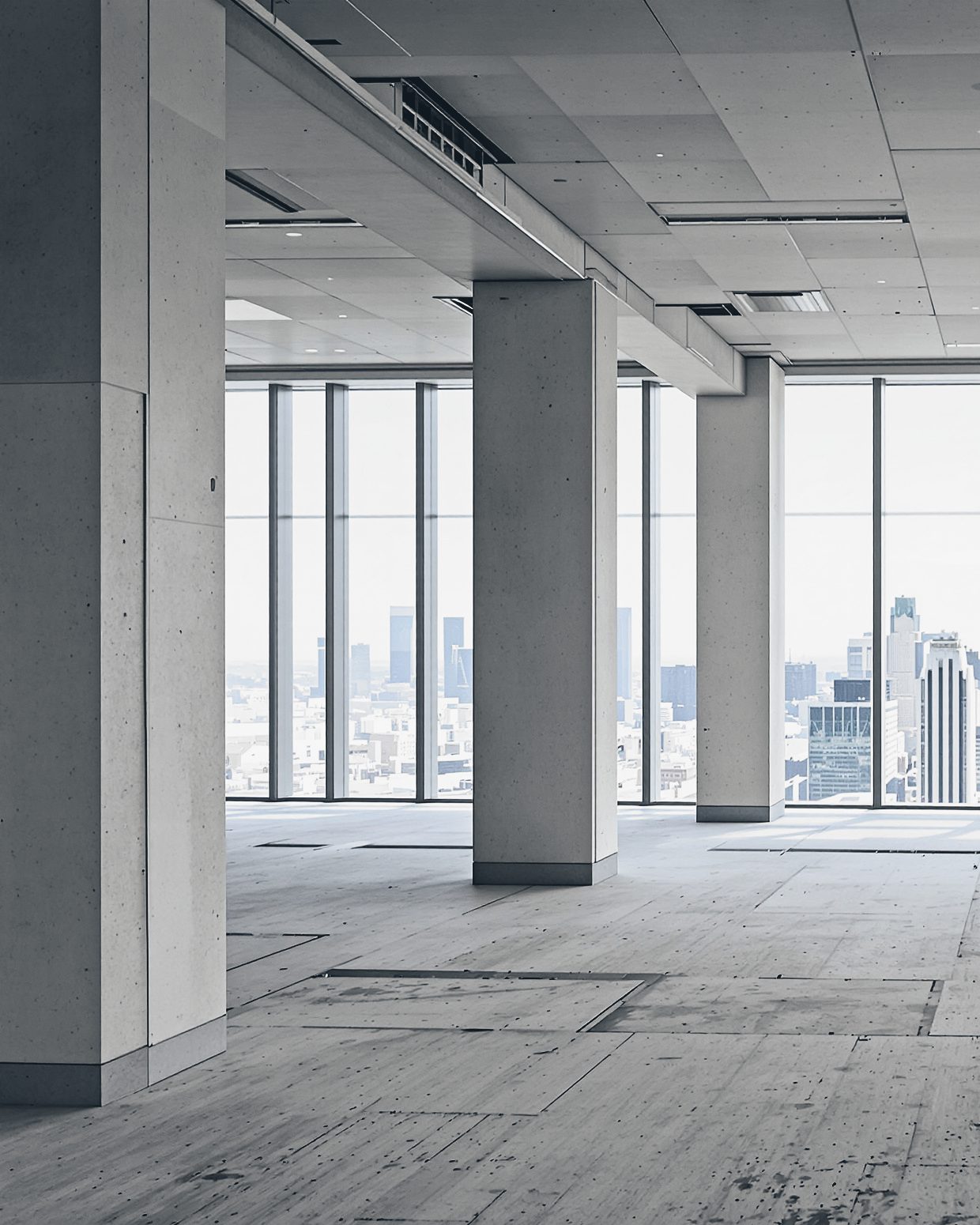 An empty upper floor of an office building: a concrete column beside floor-to-ceiling glazing and a hazy skyline.