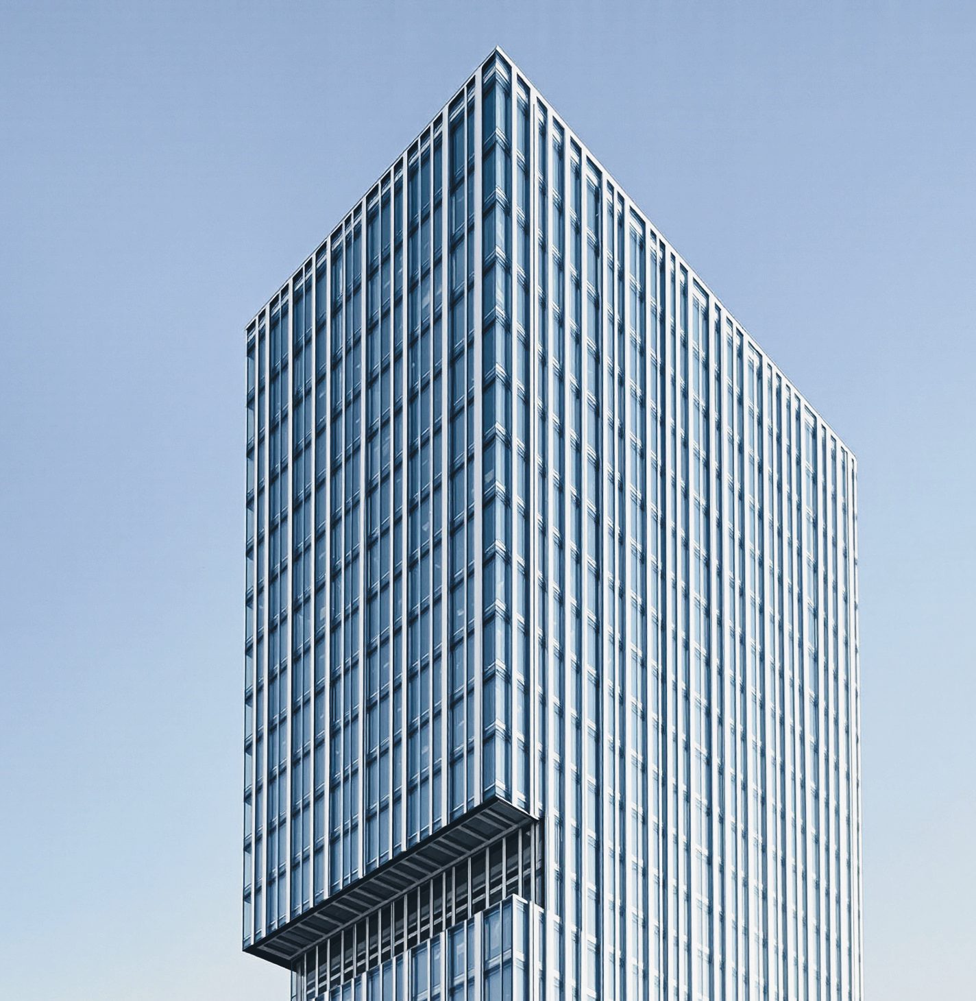A glass office tower with a cantilevered upper block and flush curtain wall, seen from a distance against a pale blue sky.